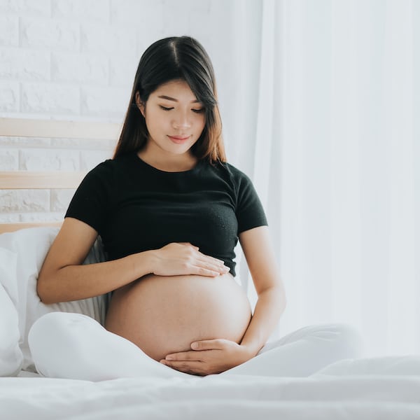 Happy Asian pregnant woman touching her belly with care in bedroom.